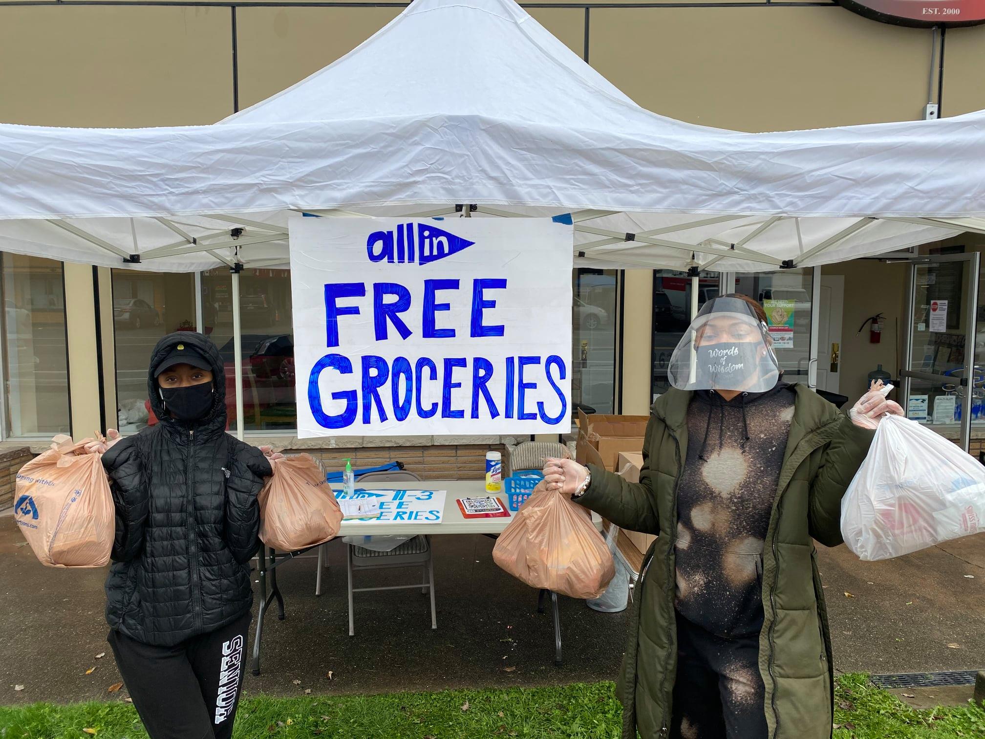 Two young Black women wearing puffy coats and face masks stand holding plastic bags of groceries outside booth with sign that says "All In: Free Groceries"